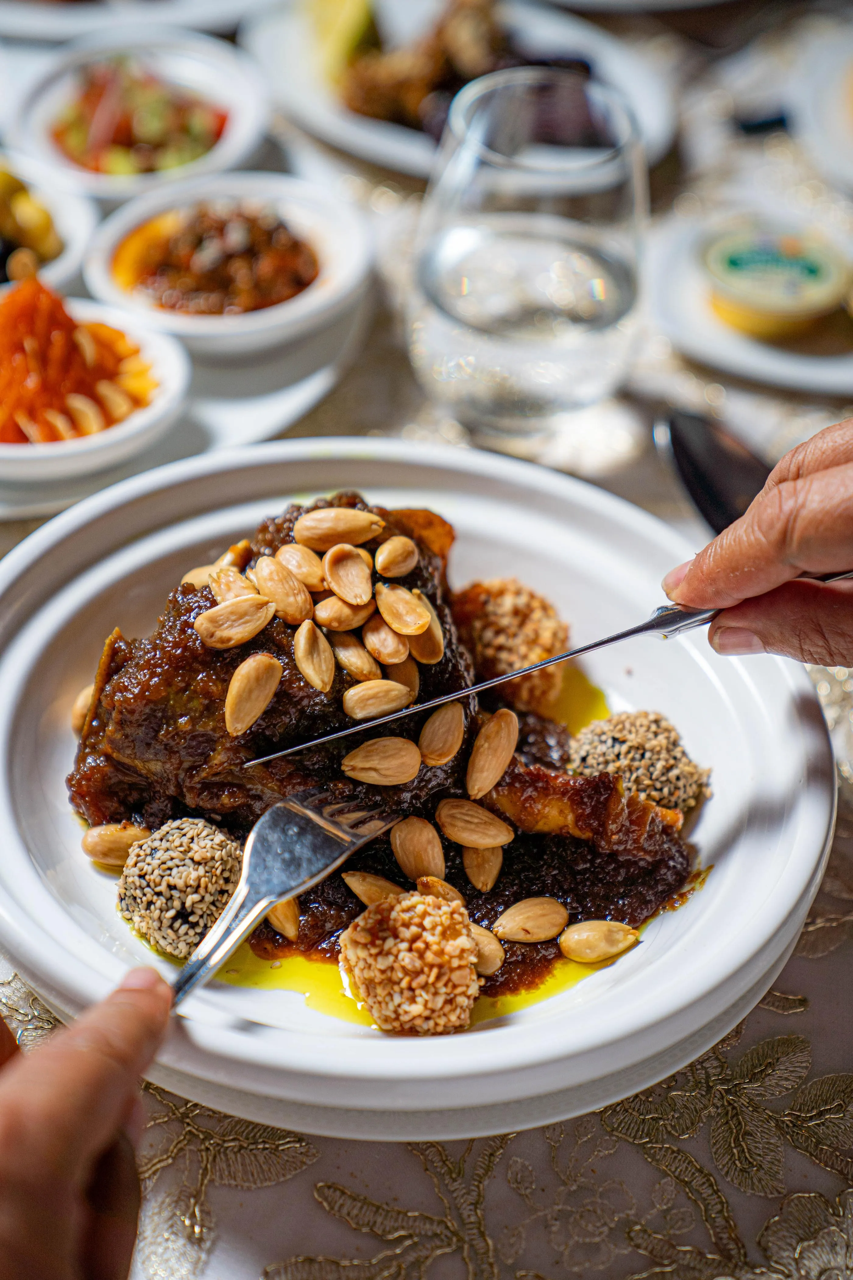 A person serves a dish with pieces of meat, almonds, and sesame seeds on a table set with various small plates.