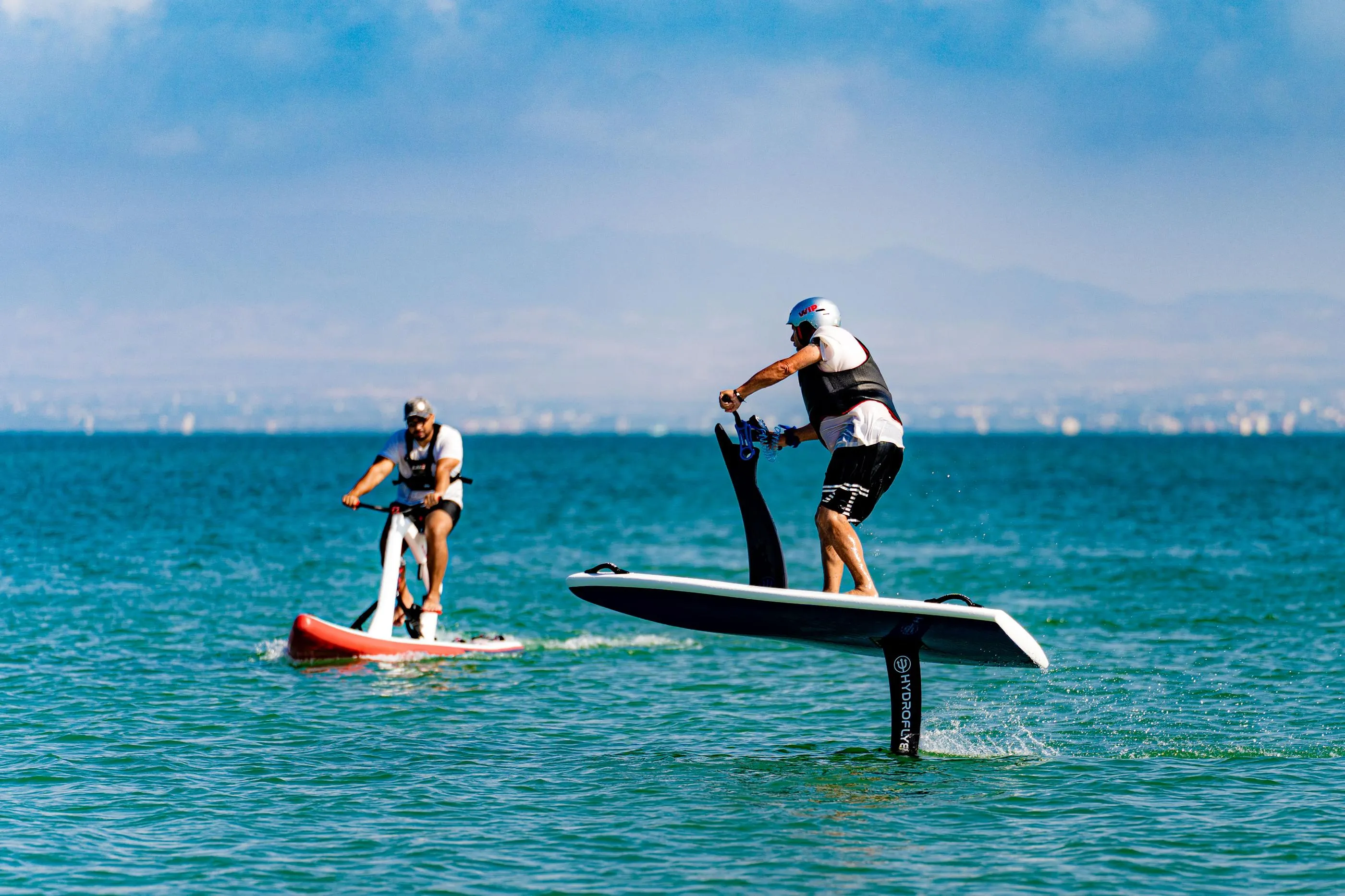 Dos personas hacen paddleboard en una extensión de agua con un fondo de ciudad lejana.