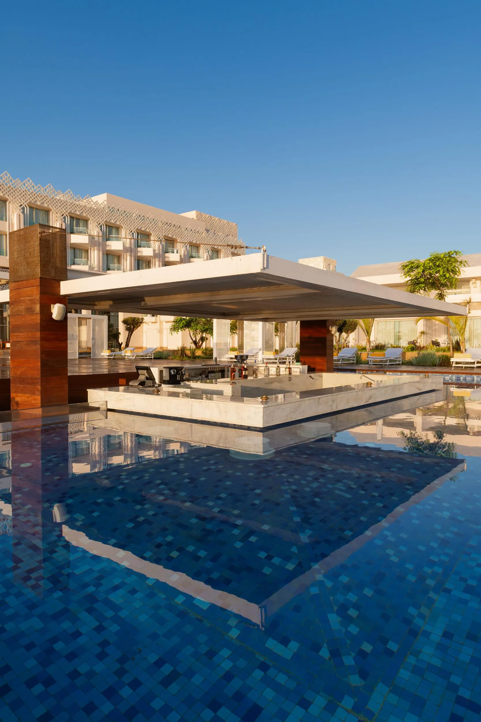 Pool with a central bar and sun loungers around, reflecting the buildings and the clear blue sky