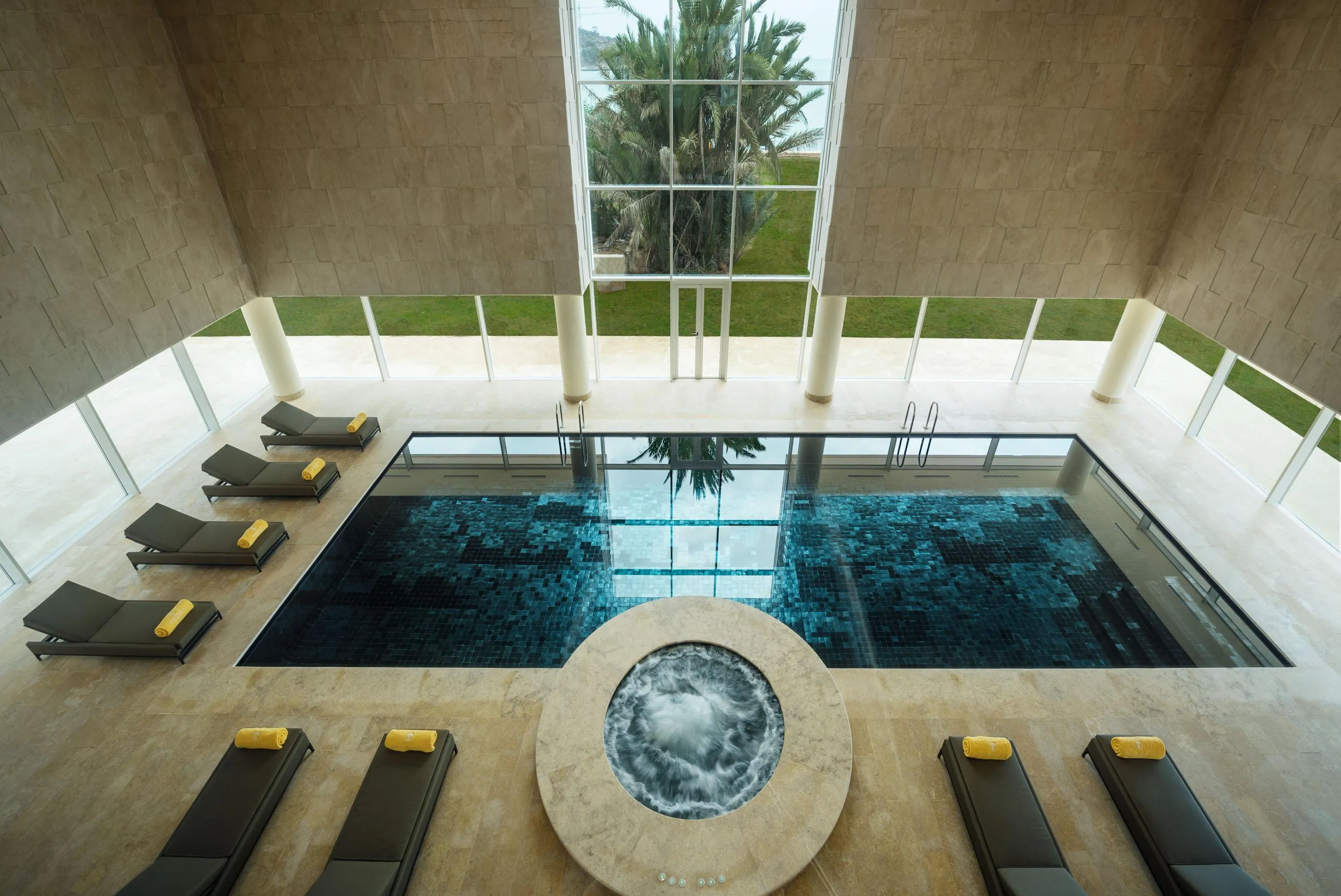 Indoor pool with sun loungers around, viewed from the ceiling with a large window and columns