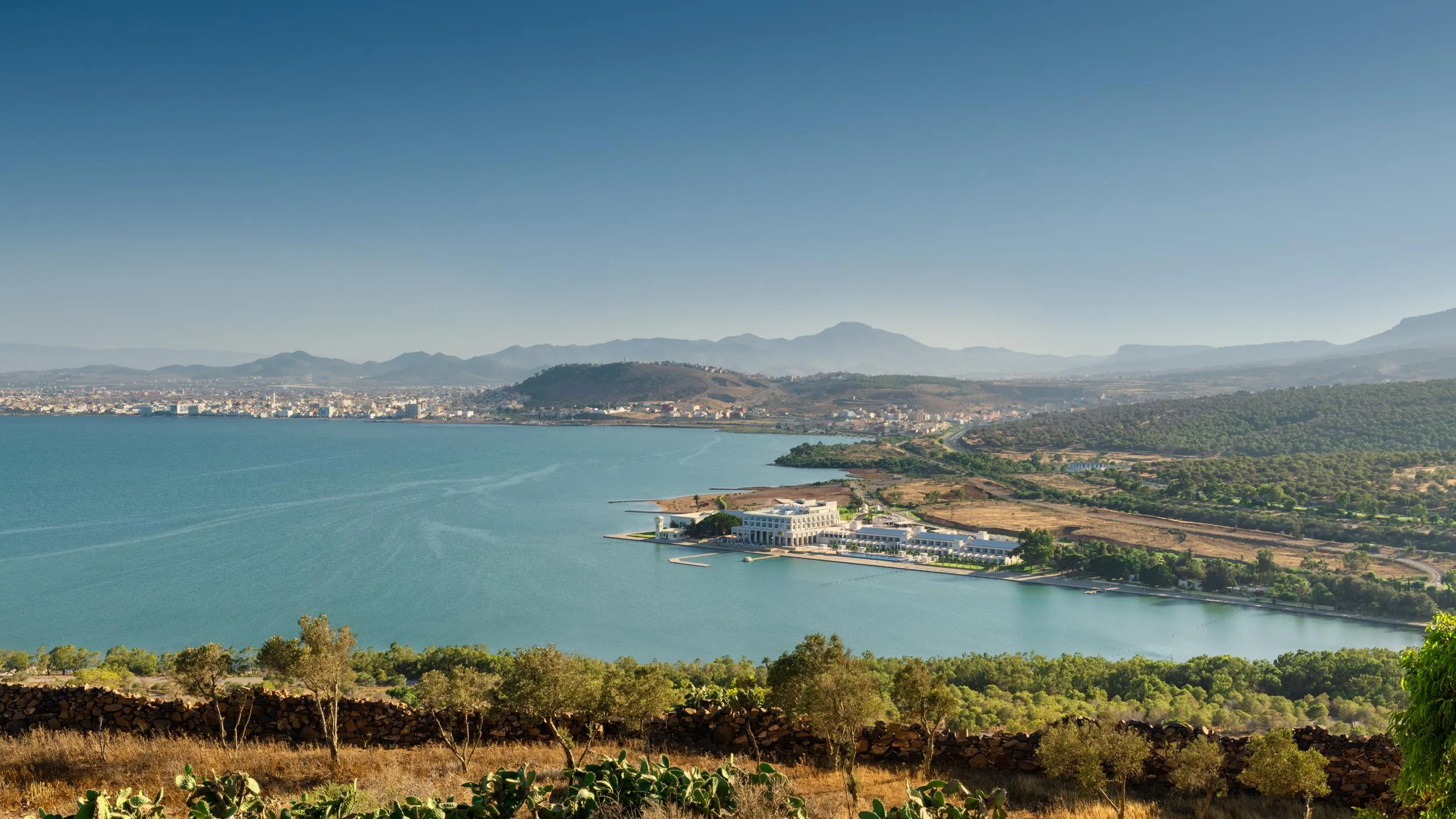 Vista panorámica de un lago rodeado de colinas y montañas con un edificio blanco en el borde de la orilla