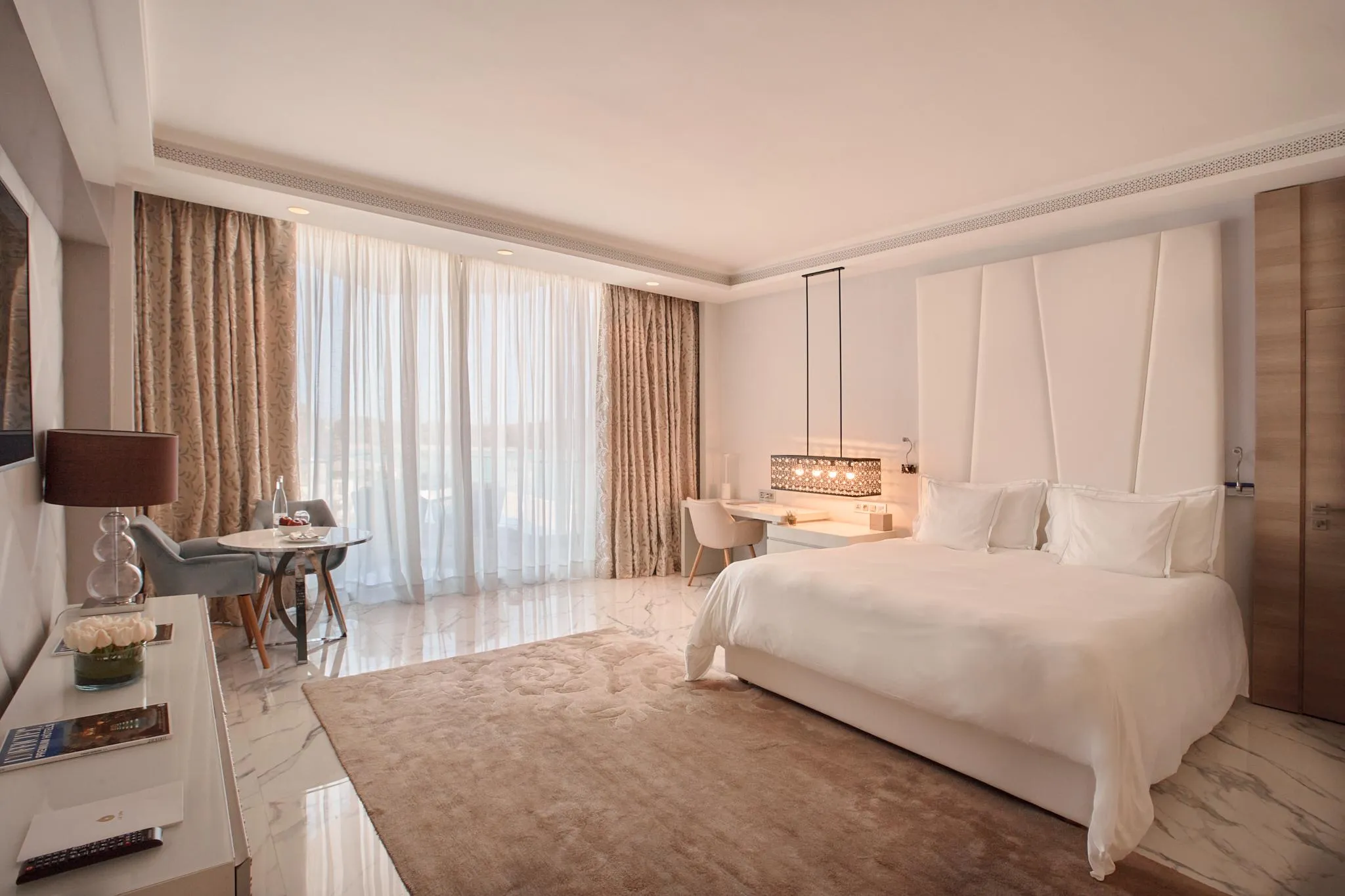 Hotel room with a large white bed, a bedside table with a lamp, a seating area near a large window with open curtains.