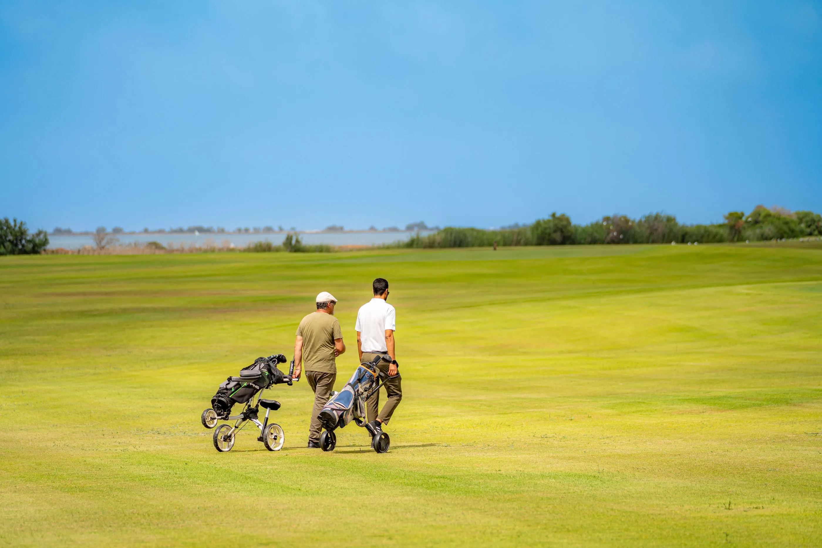 Two people walk on a golf course with club carts under a clear sky.