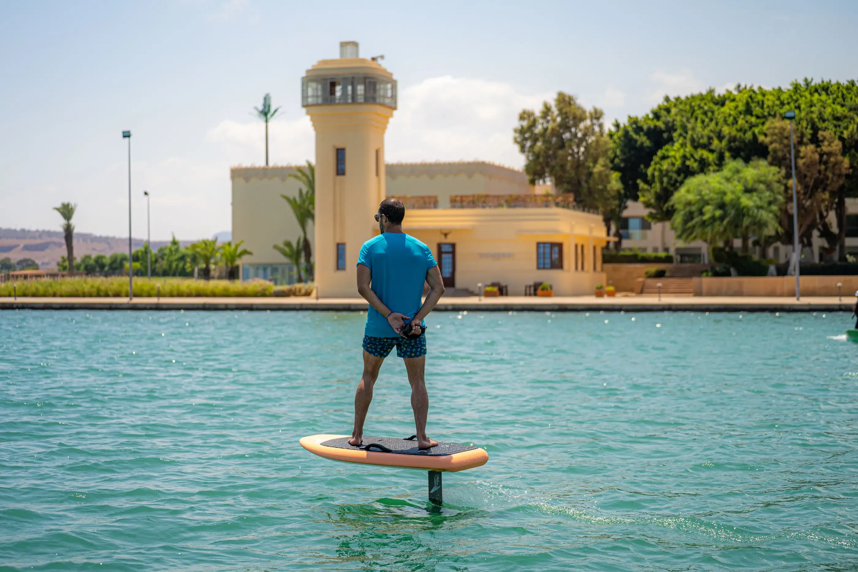 Un hombre de pie sobre una tabla de paddle en una extensión de agua, con un edificio amarillo de fondo