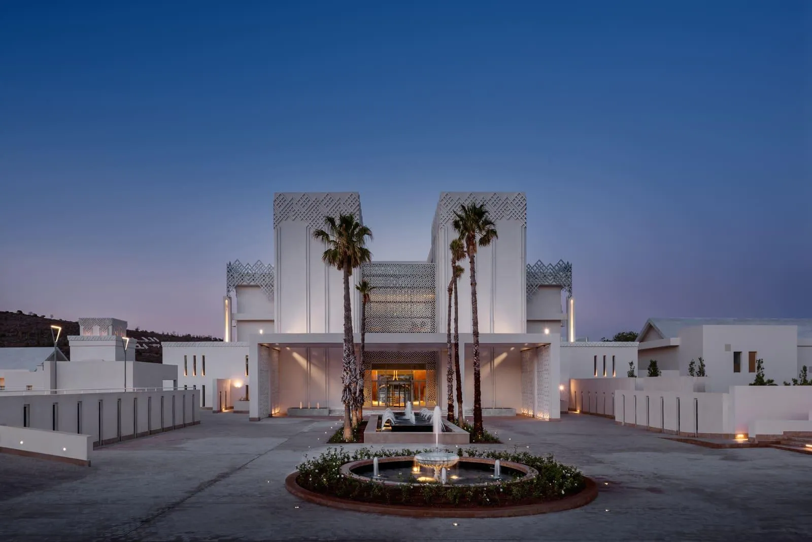 Modern building with white facade and geometric patterns, surrounded by palm trees and fountains, lit at nightfall