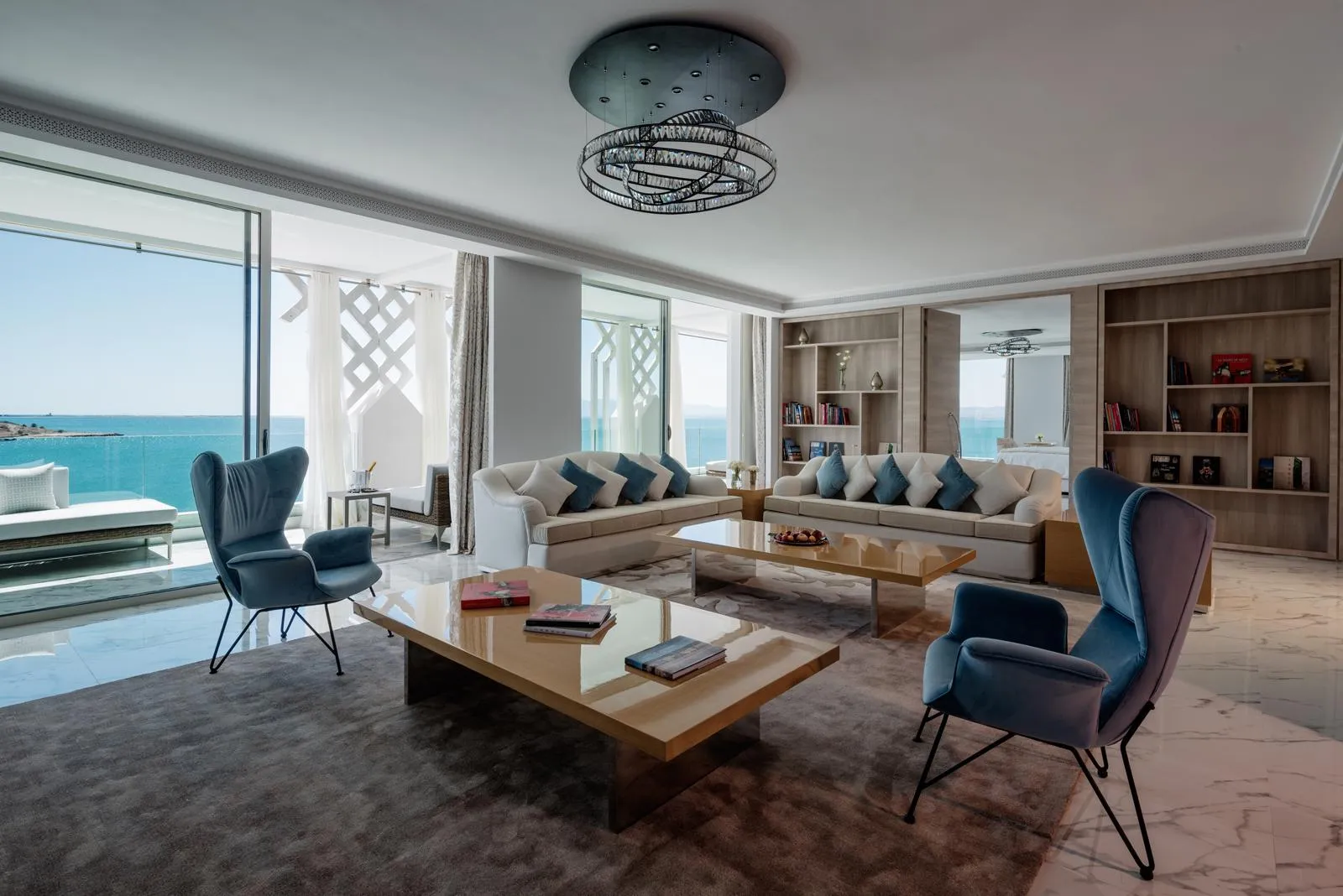 Modern living room with sea view, featuring a sofa, armchairs, a coffee table and shelves filled with books.