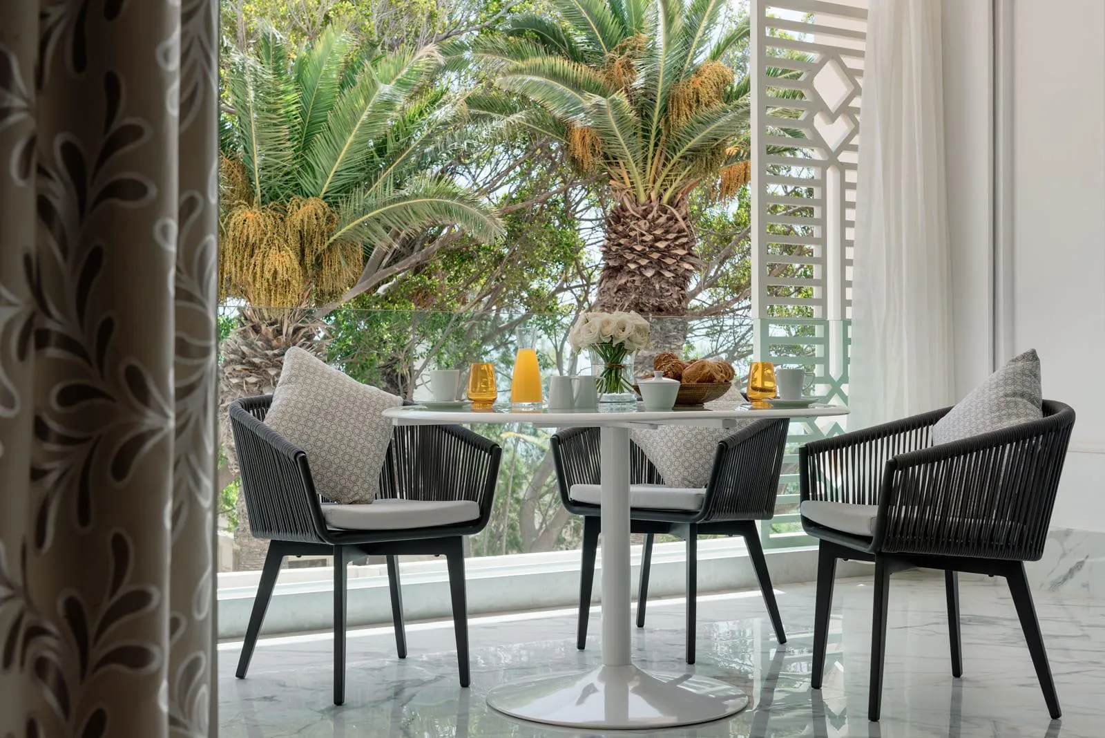 Round table with four black rattan chairs and gray cushions, seen from an open door, with plants and decorative elements on the table in the background.