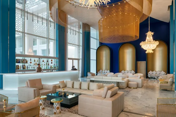 A luxurious bar and lounge with sofas, chairs, and coffee tables, lit by chandeliers and lamps, with a bartender behind the counter.