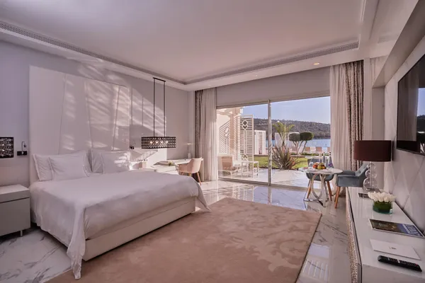 Modern hotel room with a large bed, a table and chairs near a glass door opening onto a terrace with a water view.