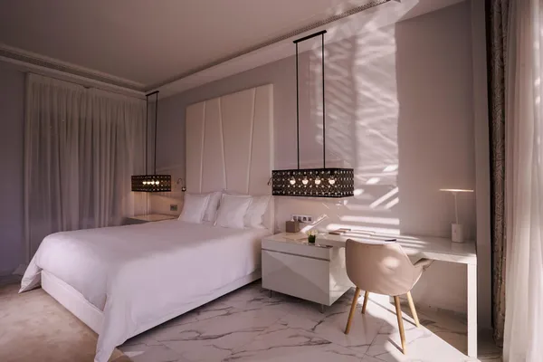 Room with a white double bed, a bedside table, and a chair near a window with translucent curtains.