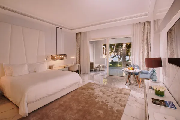 Luxurious hotel room with a large bed, a table with fruit and water bottles, and a sliding door leading to a terrace with a view of palm trees.