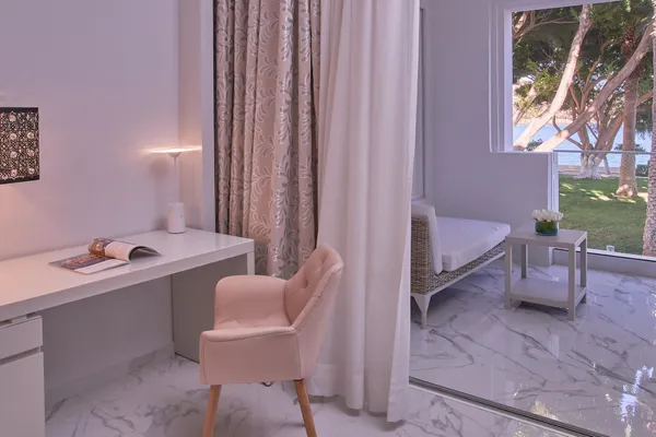 White desk with pink chair, lit lamp, magazines, and floral decoration near a window overlooking a garden with trees.