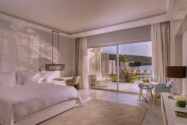 Luxurious room with white bed, work desk, and view of a terrace and outdoor landscape through large windows.