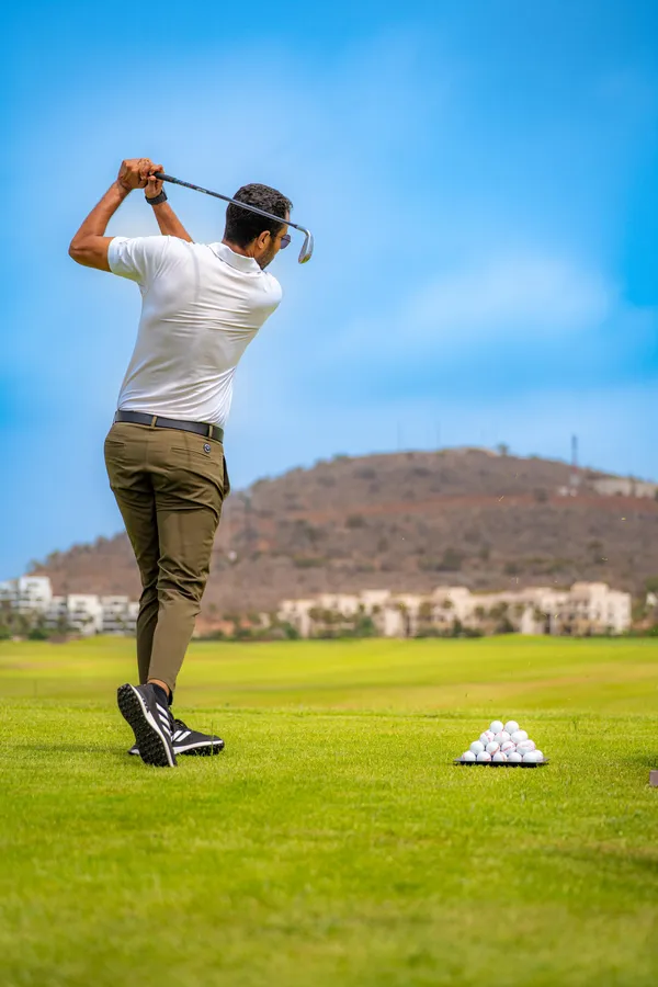 Un homme en polo blanc et pantalon vert frappe une balle de golf sur un terrain avec des balles et un tapis de départ visibles au premier plan