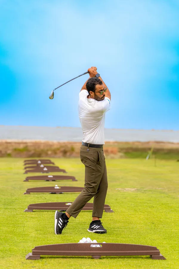 Un homme en polo blanc et pantalon vert frappe des balles de golf sur un practice en extérieur