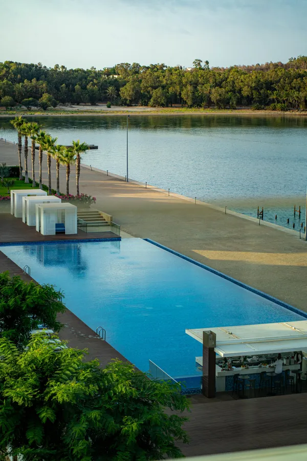 Piscine rectangulaire avec un bar et des cabanes blanches, bordée de palmiers, face à un lac et une plage de sable