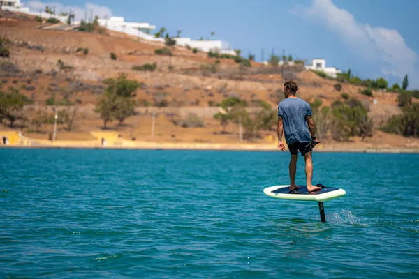 A man standing on an electric paddleboard on a body of water with an arid landscape in the background.