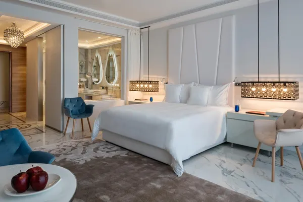 Luxurious room with white bed, two armchairs, bedside table, and view of a bathroom with oval mirrors.