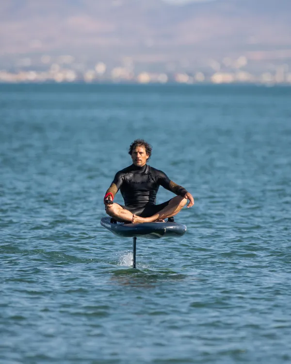 Un homme en combinaison de surf est assis en position méditative sur une planche flottant sur l'eau, avec des lumières au loin à l'horizon