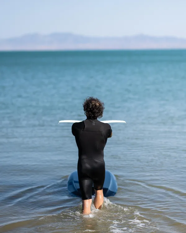 Une personne en combinaison noire se tient dans l'eau peu profonde, face à la mer, tenant une planche de surf blanche