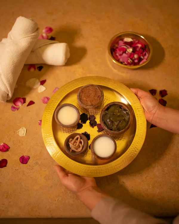 A yellow tray with several small containers holding various substances is held by two hands, with rose petals scattered around.