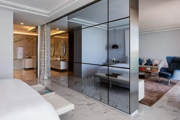 Modern interior with a mirror separating a bedroom and a living room, featuring white furniture and a seating area with a sofa and armchair.
