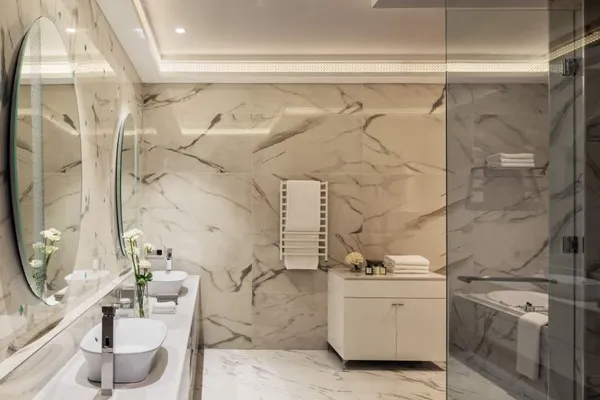 Modern bathroom with double sink, round mirror, glass shower, and beige marble walls