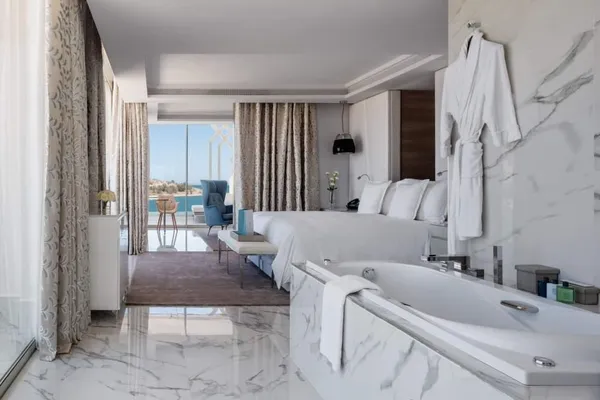 Luxurious room with white bed, bathtub, and view of a terrace with blue chair and distant sea.